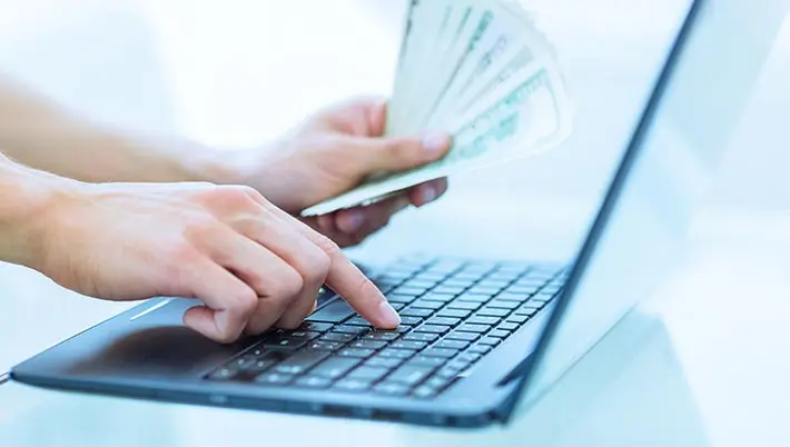 hand holding dollar bills while typing on a laptop  