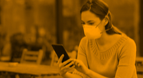 woman sitting at a coffee shop wearing a mask and looking at her phone